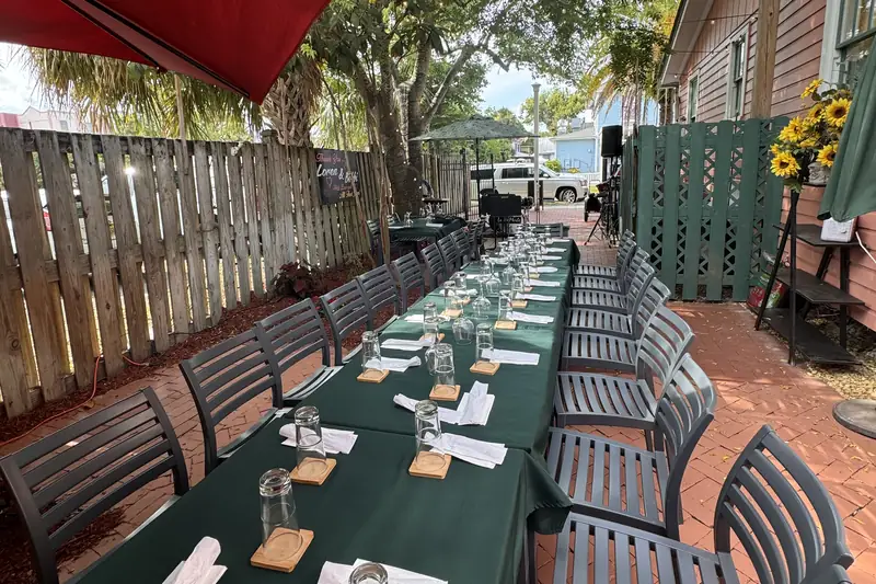 Courtyard set for a private party: long tables with green tablecloths under string lights and shade trees