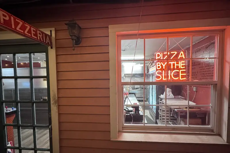 Pizza Street Kitchen at night: a glowing red "Pizza by the Slice" neon sign in the window next to the Pizzeria door